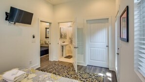 Habitación de hotel con cama, televisor montado en la pared, baño con lavamanos y espejo, y una puerta que da a otra habitación.