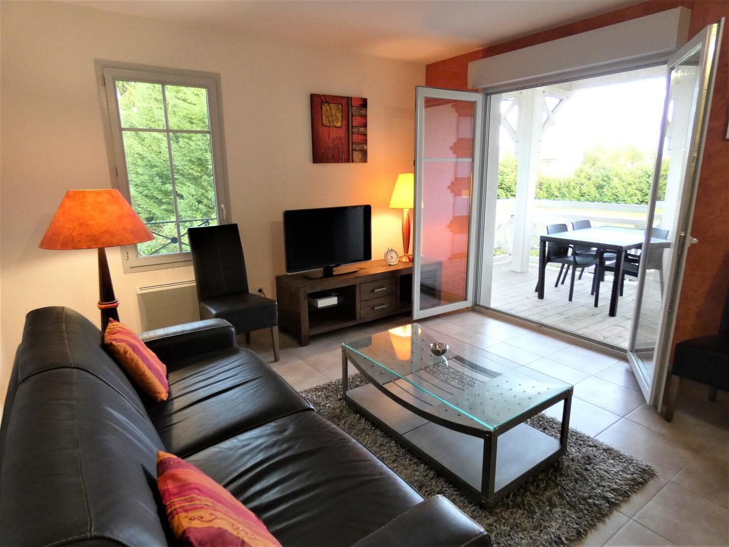 A living room with a sofa, TV, and a glass coffee table. There's a sliding glass door leading to an outdoor patio with a table and chairs.