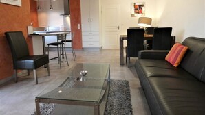 A modern living room with a glass coffee table, black leather sofa, and a dining area with a table and chairs. - Chez Detché - Mimizan (MIMIZAN)