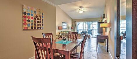 A dining area with a wooden table and chairs, a ceiling fan, and a view of the kitchen and living area through large windows.