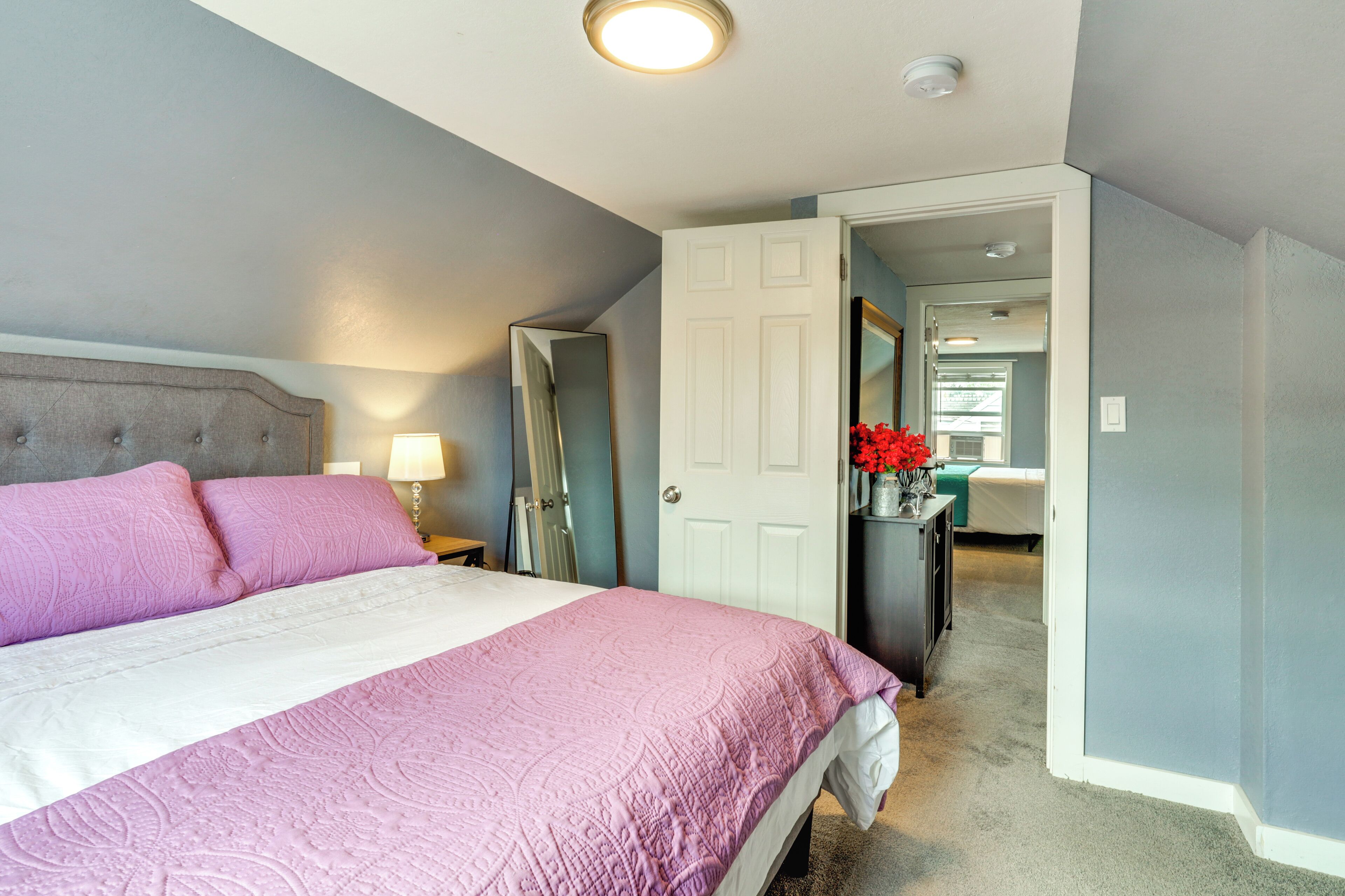 A bedroom with a bed, a nightstand, a lamp, a mirror, and a door leading to another room.