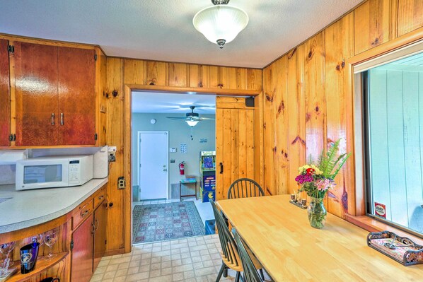 A kitchen with wooden cabinets, a microwave, a dining table with chairs, a vase with flowers, and a view through an open doorway to another room.