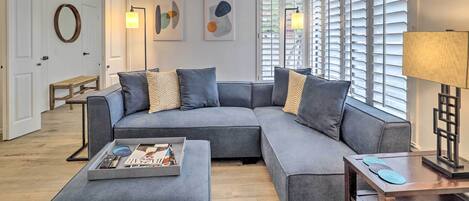 Un salon moderne avec un canapé d’angle gris, une table basse avec des magazines et une lampe sur une table d’appoint.