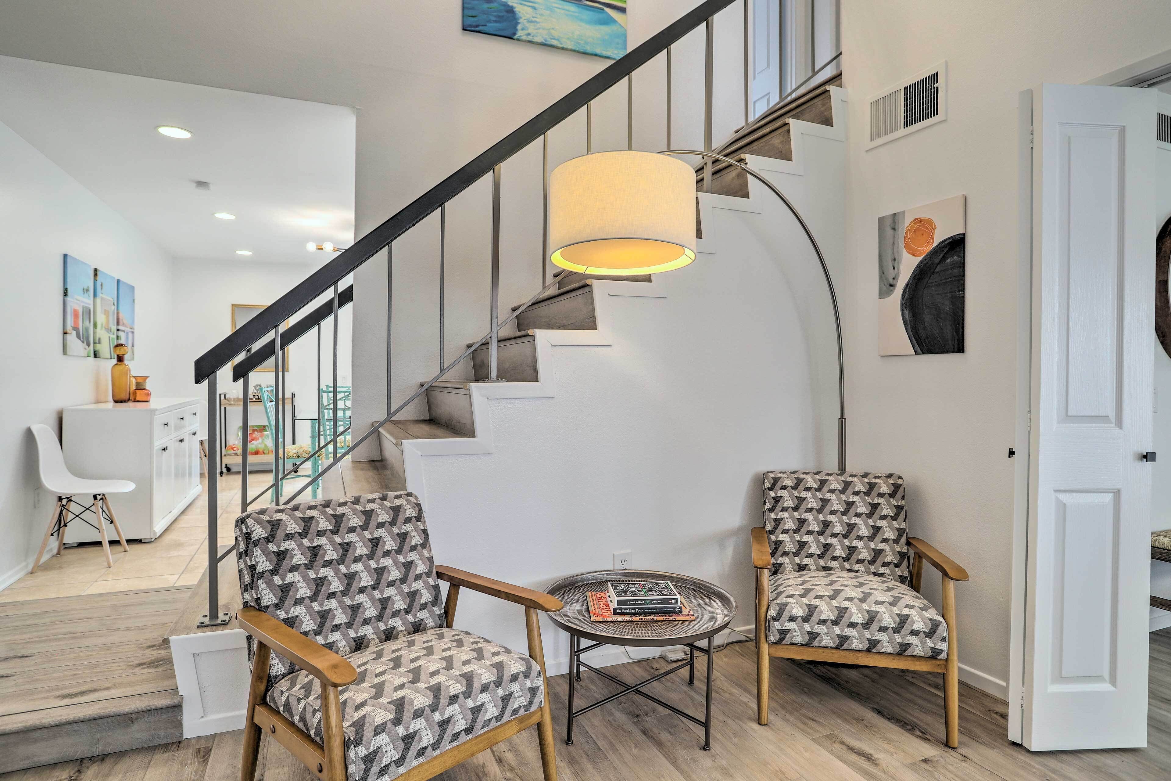 A modern living room with a staircase, two patterned armchairs, a small round table with books, and a large floor lamp.