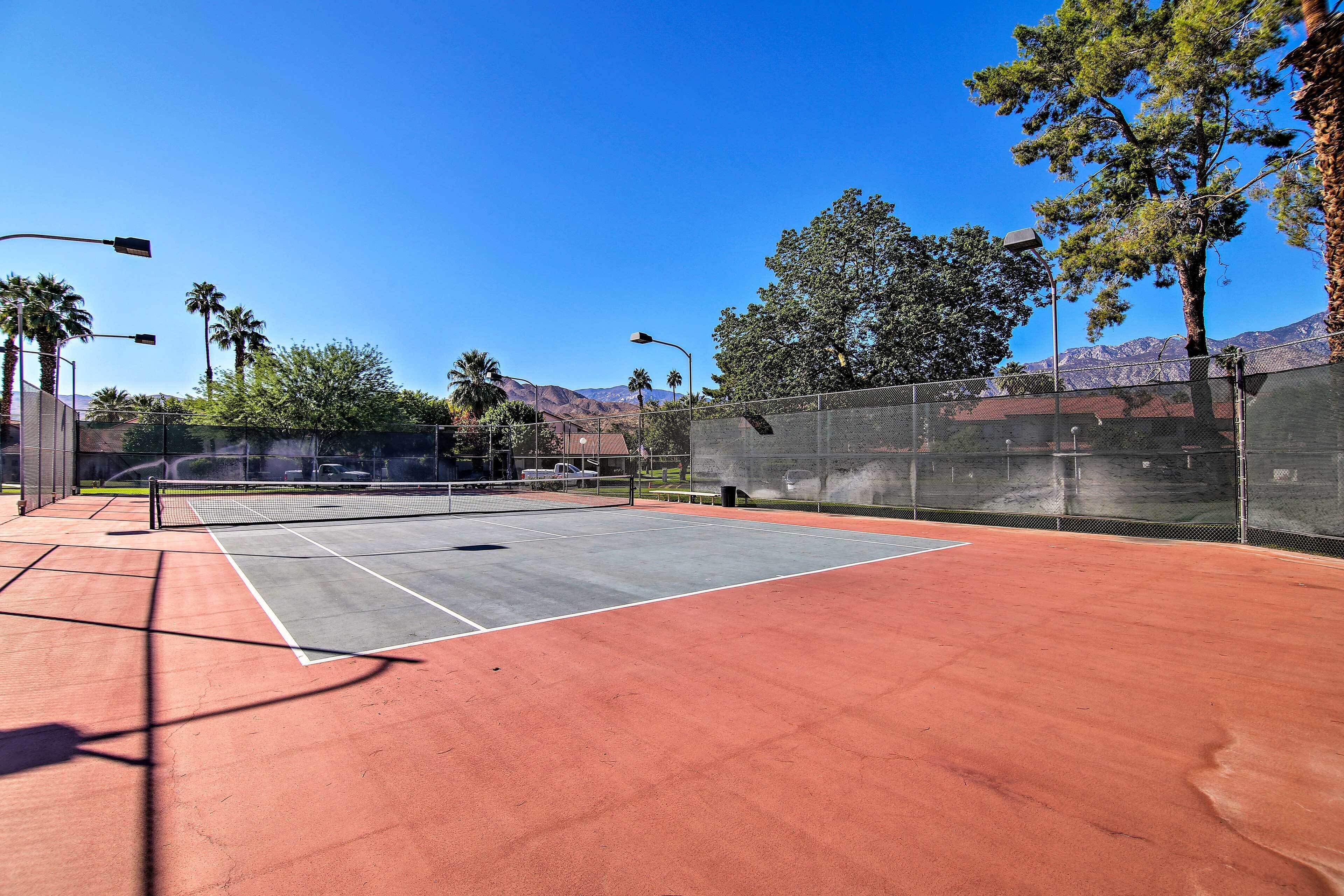 A tennis court with a net, surrounded by a fence, palm trees, and mountains in the background.