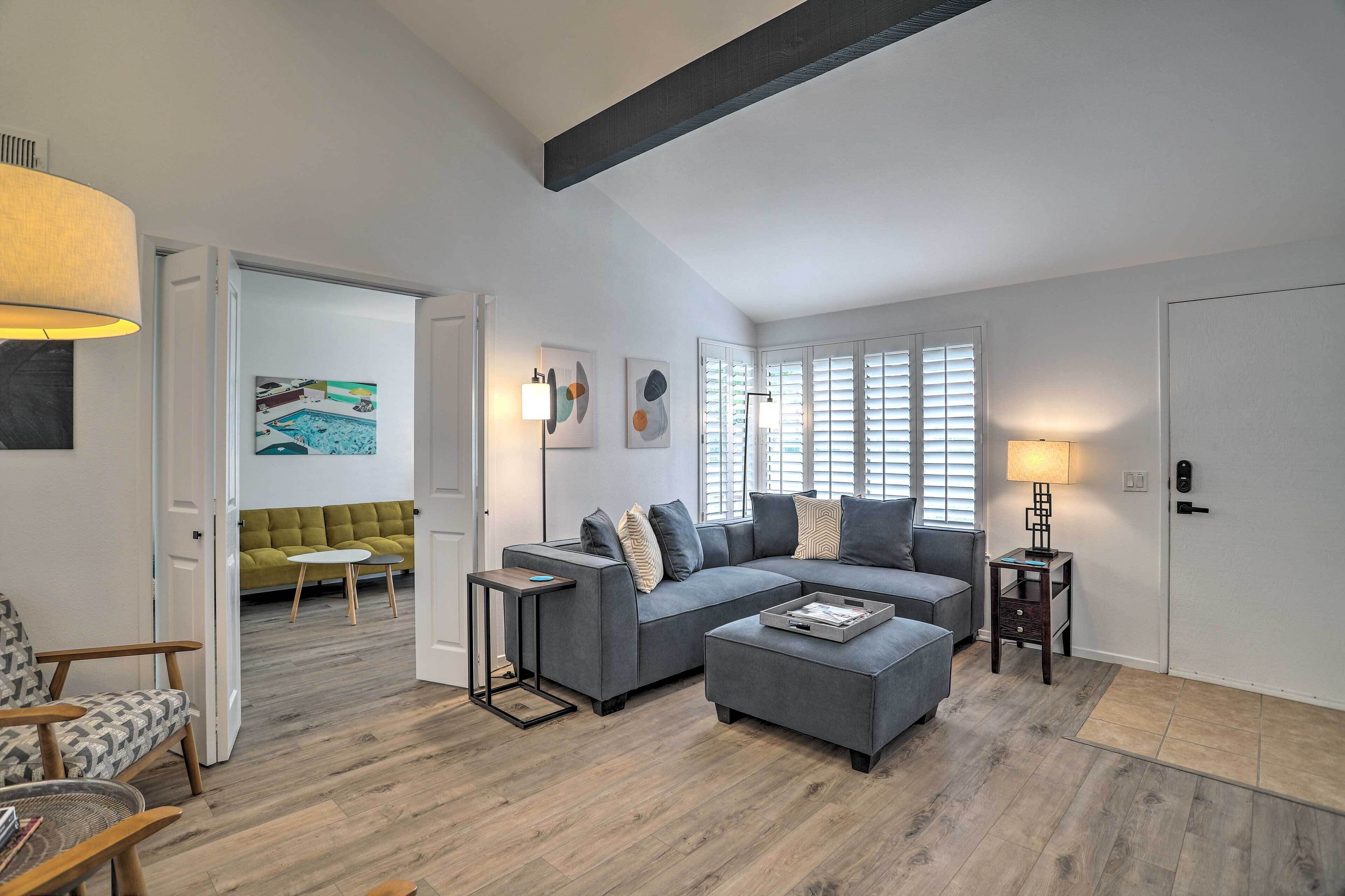 A modern living room with a grey sectional sofa, a small table, and a lamp. There is a door leading to another room with a green sofa and a painting.