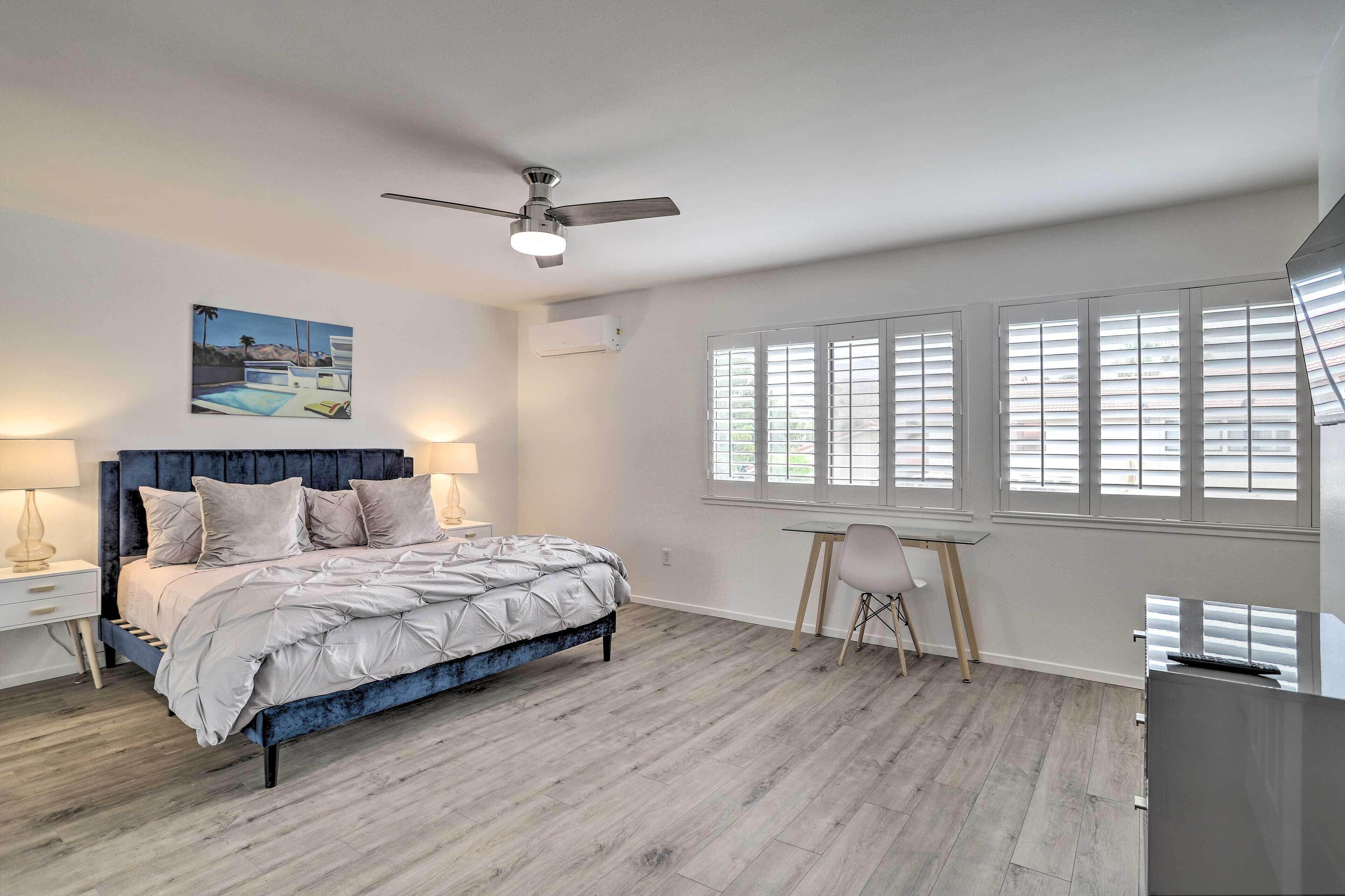 Ein Schlafzimmer mit Bett, Schreibtisch und Stuhl. An der Decke hängt ein Ventilator, an der Wand hängt ein Bild und es gibt ein Fenster mit Jalousien.