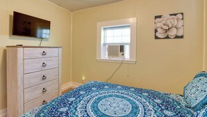 Un dormitorio con cama, cómoda, televisor montado en la pared y ventana con aire acondicionado.