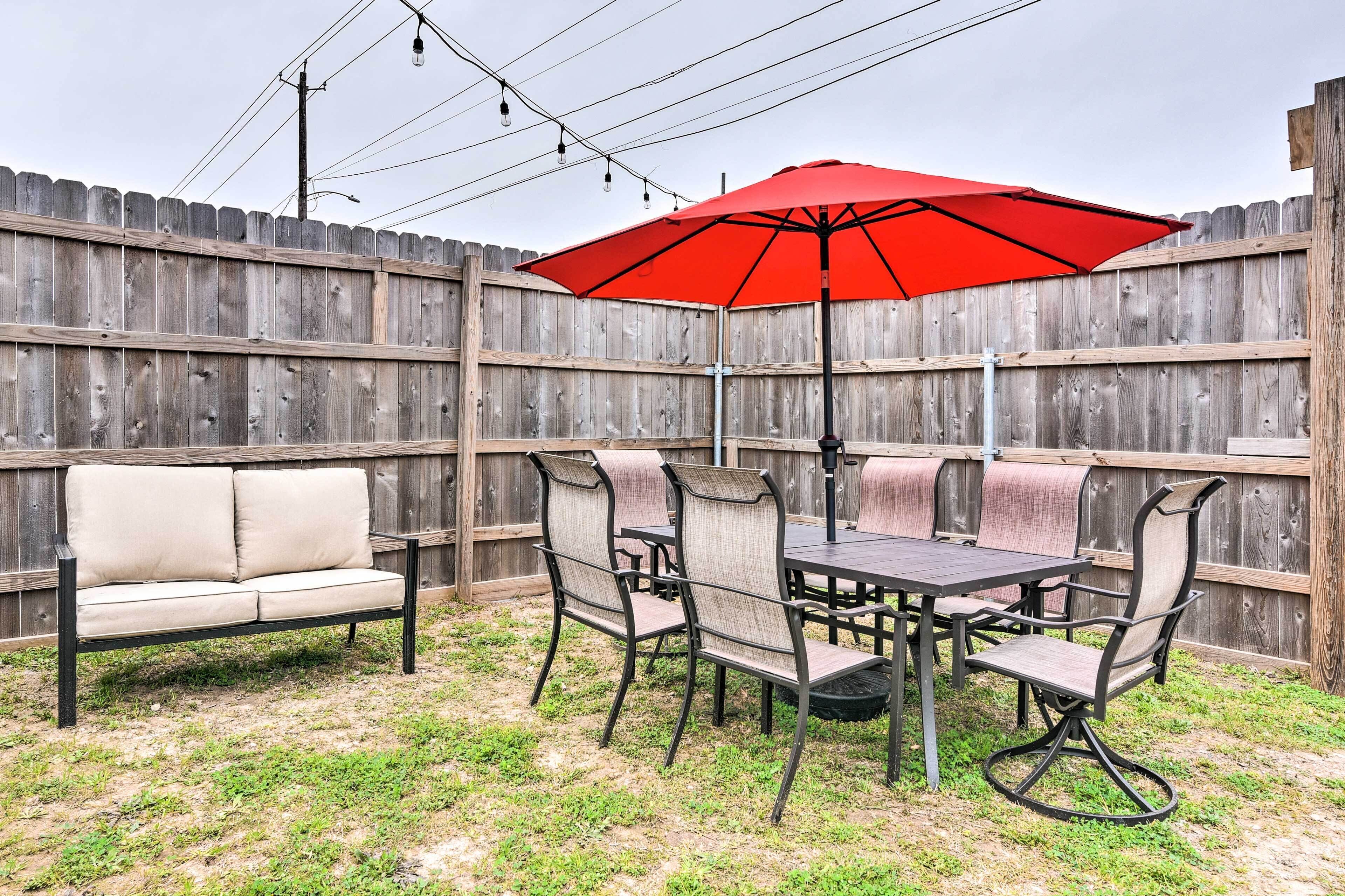 A backyard patio with a wooden fence, a beige sofa, a dining table with chairs, and a red umbrella.