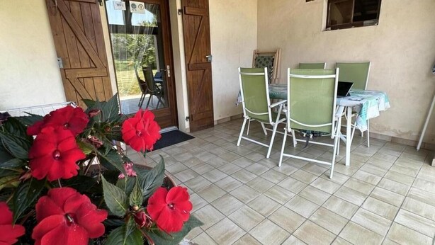 A covered patio with a table and chairs, a wooden door, and potted red flowers. - Le Tastet - Saint LON LES Mines (SAINT LON LES MINES)