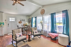 A cozy living room with wicker furniture, a dining table, and a large window with blue curtains. - Lively Galveston Home w/ Deck & Beach Views! (Galveston)