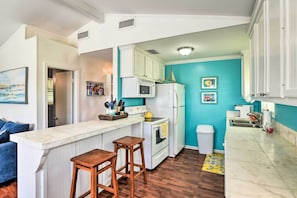 A kitchen with a white countertop, wooden stools, a stove, a microwave, and a refrigerator. There is a trash can and a sink. - Lively Galveston Home w/ Deck & Beach Views! (Galveston)