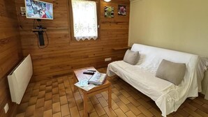 A cozy room with a wooden interior, a white sofa, a small table with a book, a TV mounted on the wall, and a window with lace curtains. - Les Ajoncs - Brocas - Gîte (BROCAS)
