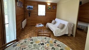 A cozy room with wooden walls, a white sofa, a table with a floral tablecloth, and a window with a curtain. - Les Ajoncs - Brocas - Gîte (BROCAS)