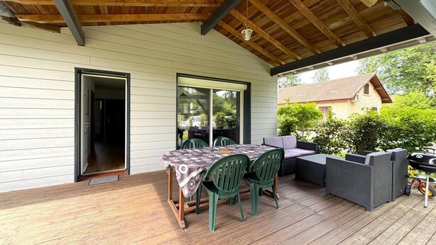 En udendørs terrasse med et bord og stole, et overdækket siddeområde og udsigt til et hus i baggrunden.