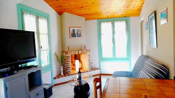 A cozy living room with a wooden ceiling, a television on a cabinet, a fireplace, and a dining area with a table and chairs. - Le Gîte du Pont Blanc (ST-JULIEN-EN-BORN)