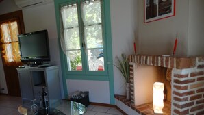 A living room with a television, a glass table, a fireplace, and a window with curtains. - Le Gîte du Pont Blanc (ST-JULIEN-EN-BORN)