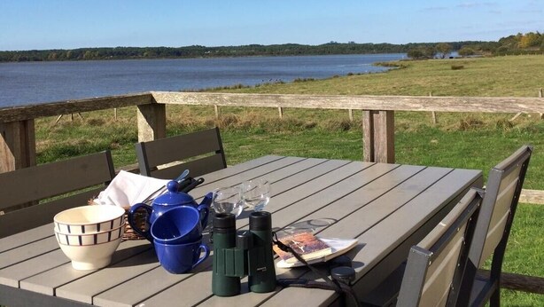 A wooden table with chairs, a bowl, a mug, binoculars, and a book, set against a backdrop of a lake and grassy area. - Lacoste 2 - ORX - Gîte (ORX)