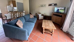 A living room with a blue sofa, a wooden coffee table, a television, and a kitchen area in the background. - Lacoste 2 - ORX - Gîte (ORX)