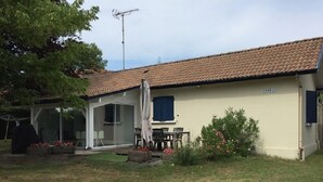 A single-story house with a tiled roof, a covered patio with a table and chairs, and a small garden area with flowers and shrubs. - Erable - Ste-eulalie-en-born (STE-EULALIE-EN-BORN)