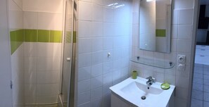 Une salle de bain avec des carreaux blancs et verts, une douche en verre, un lavabo avec un miroir et une tasse verte posée sur le comptoir.