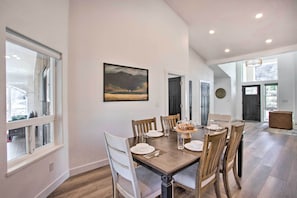 Uma sala de jantar com mesa de madeira posta para seis, cadeiras brancas e um quadro na parede.