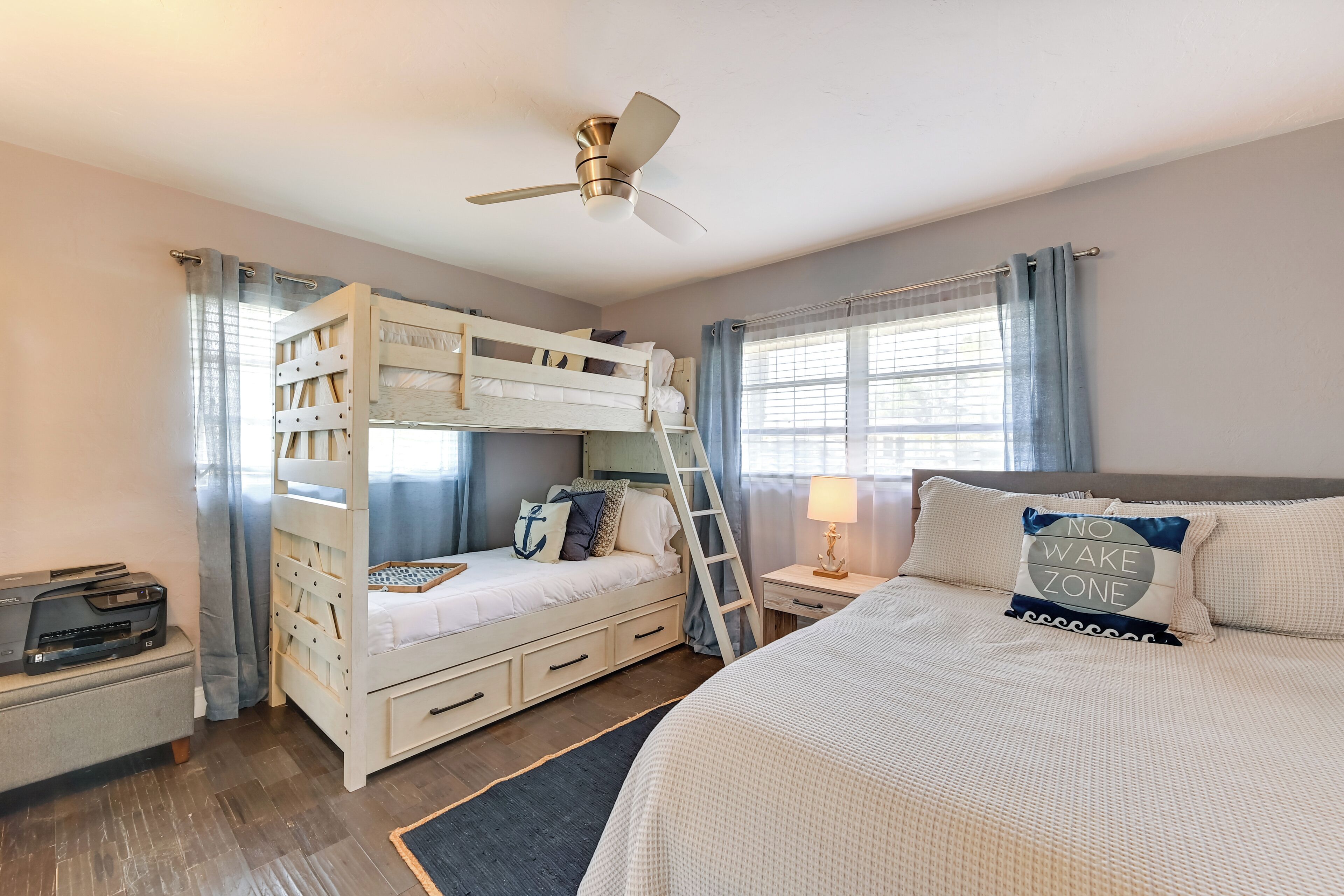 A bedroom with a bunk bed, a desk with a printer, a nightstand with a lamp, and a window with curtains.