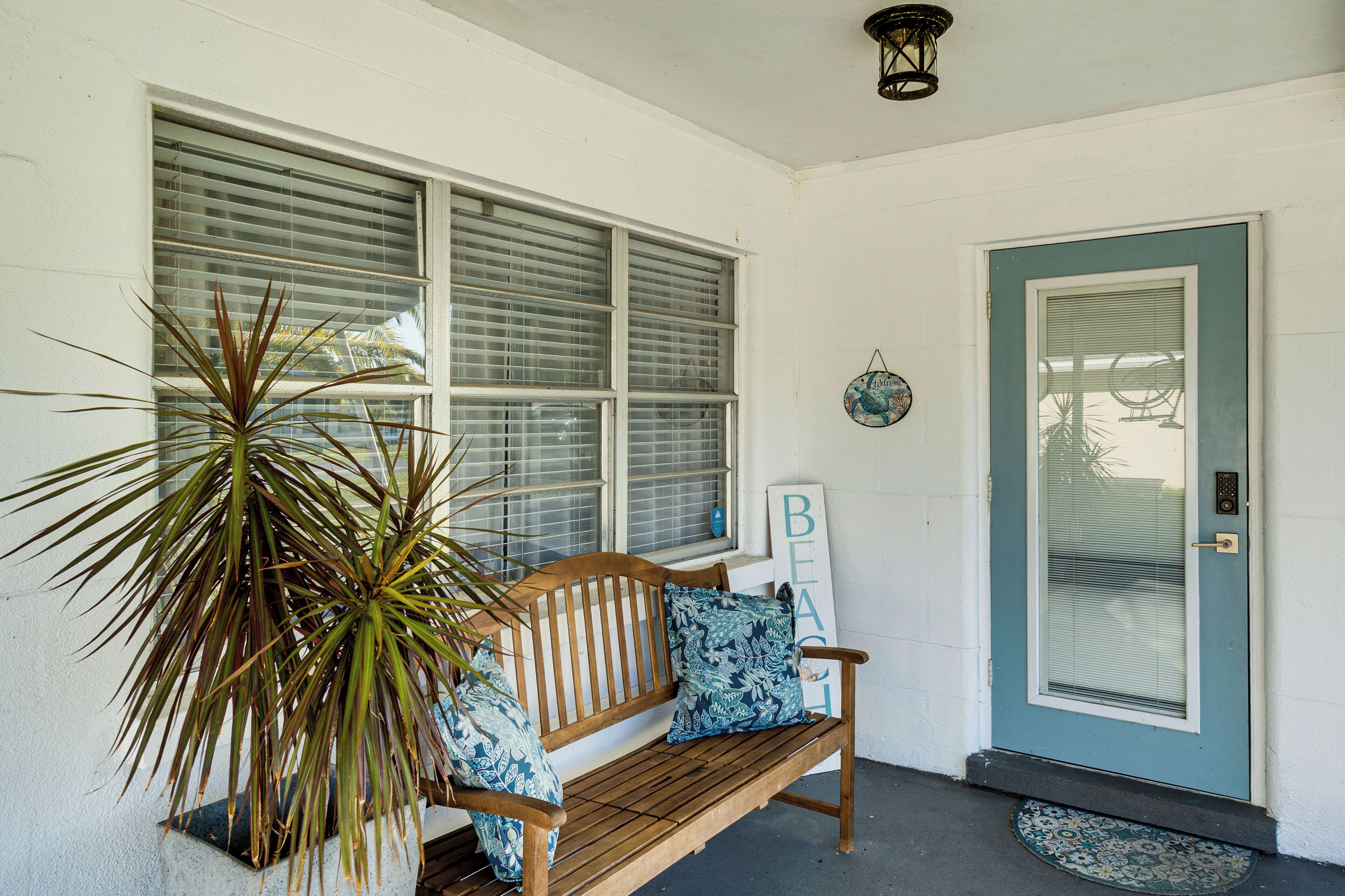 A wooden bench with blue patterned cushions, a potted plant, and a door with a sign that reads "BEACH".