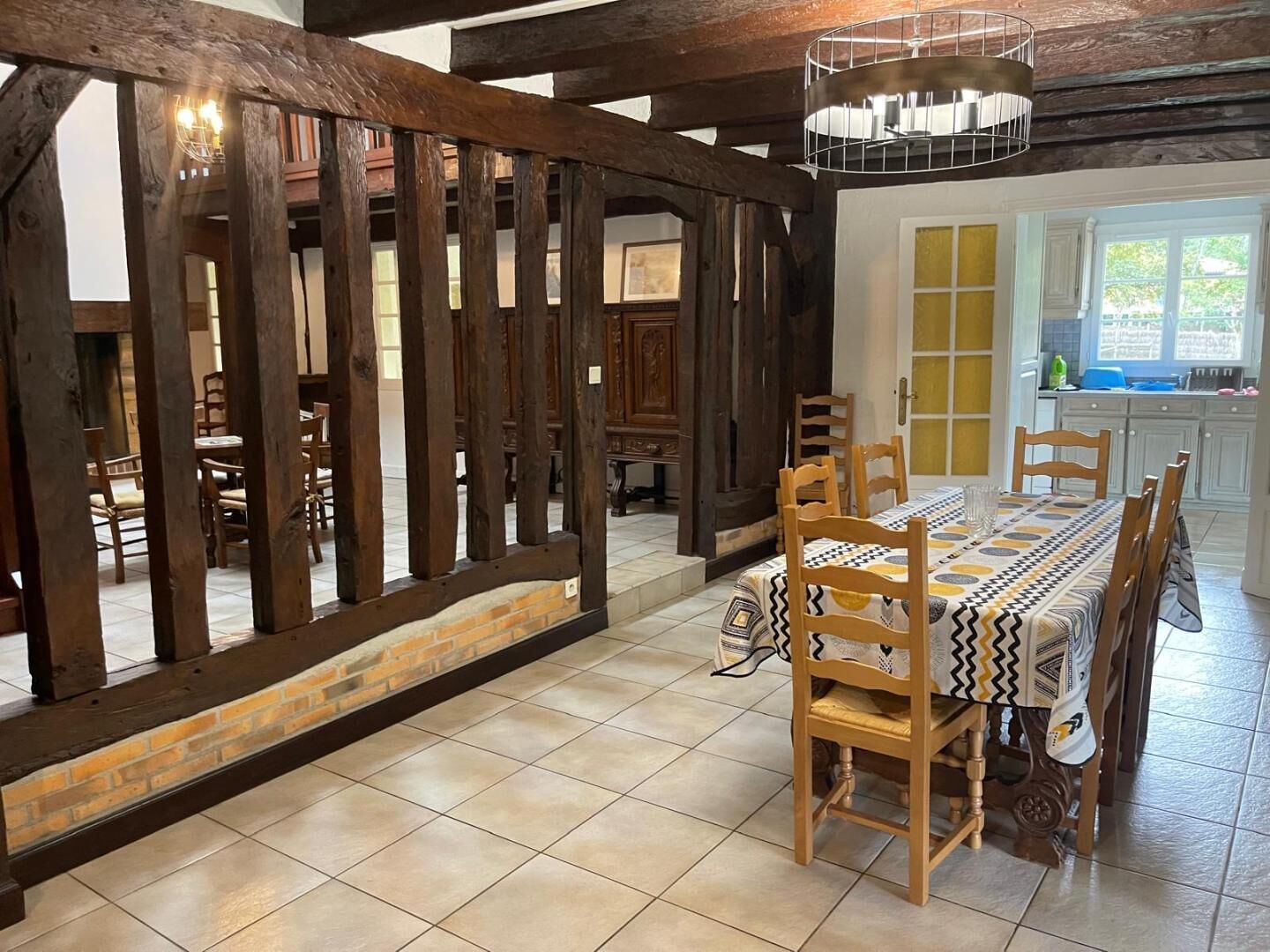 A dining area with a wooden table and chairs, a brick wall, and a view of a kitchen with a window.