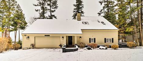 A two-story house with a garage, surrounded by snow-covered ground and tall trees.