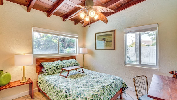 A bedroom with a bed, two windows, a ceiling fan, a table, and a chair. - Ormond Beach Retreat: Walk to Shore & Shops! (Ormond Beach)