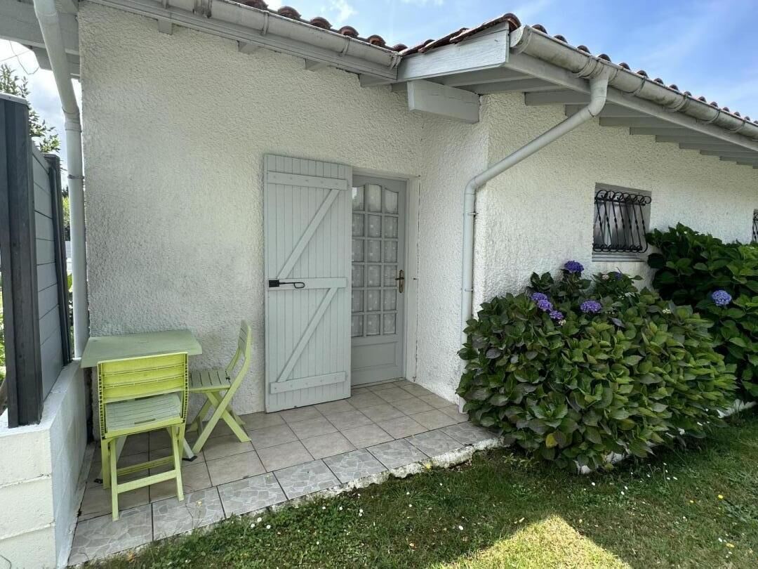 Une maison à l’extérieur blanc, avec une porte d’entrée, une petite fenêtre ornée d’une grille métallique, une table et une chaise vertes destinées à l’extérieur, et un jardin bien entretenu avec des arbustes verts et des fleurs violettes.