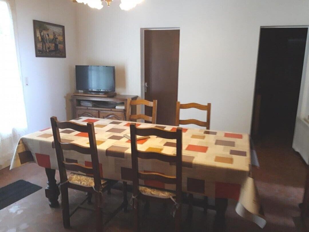 Un comedor con una mesa dispuesta para cuatro, sillas de madera, un televisor sobre un mueble bajo y una puerta al fondo.