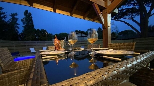 An outdoor dining area with a glass table, wicker chairs, and a view of trees and a building in the background. - Gîte de Linxe - Linxe (LINXE)