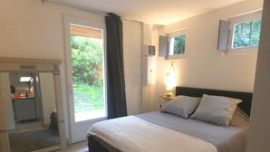 A bedroom with a bed, a nightstand with a lamp, a window with curtains, and a view of greenery outside. - Gîte de Linxe - Linxe (LINXE)