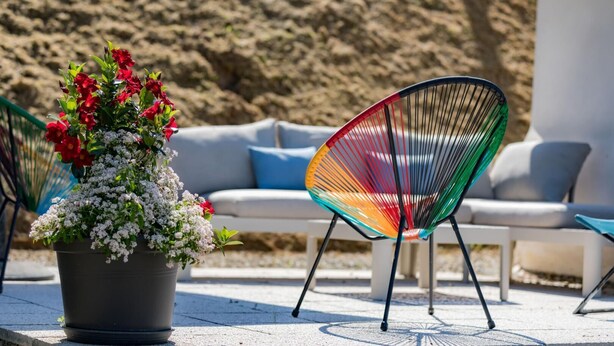 A potted plant with red and white flowers, a colorful chair, and a white sofa with blue cushions. - Gîte Capérot - Eyres Moncube (EYRES MONCUBE)