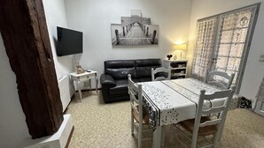 A living room with a black leather sofa, a dining table with white chairs, a TV mounted on the wall, and a painting above the sofa.