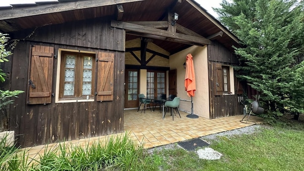 A rustic wooden building with a covered patio, a dining area with chairs, and a large tree in the background.