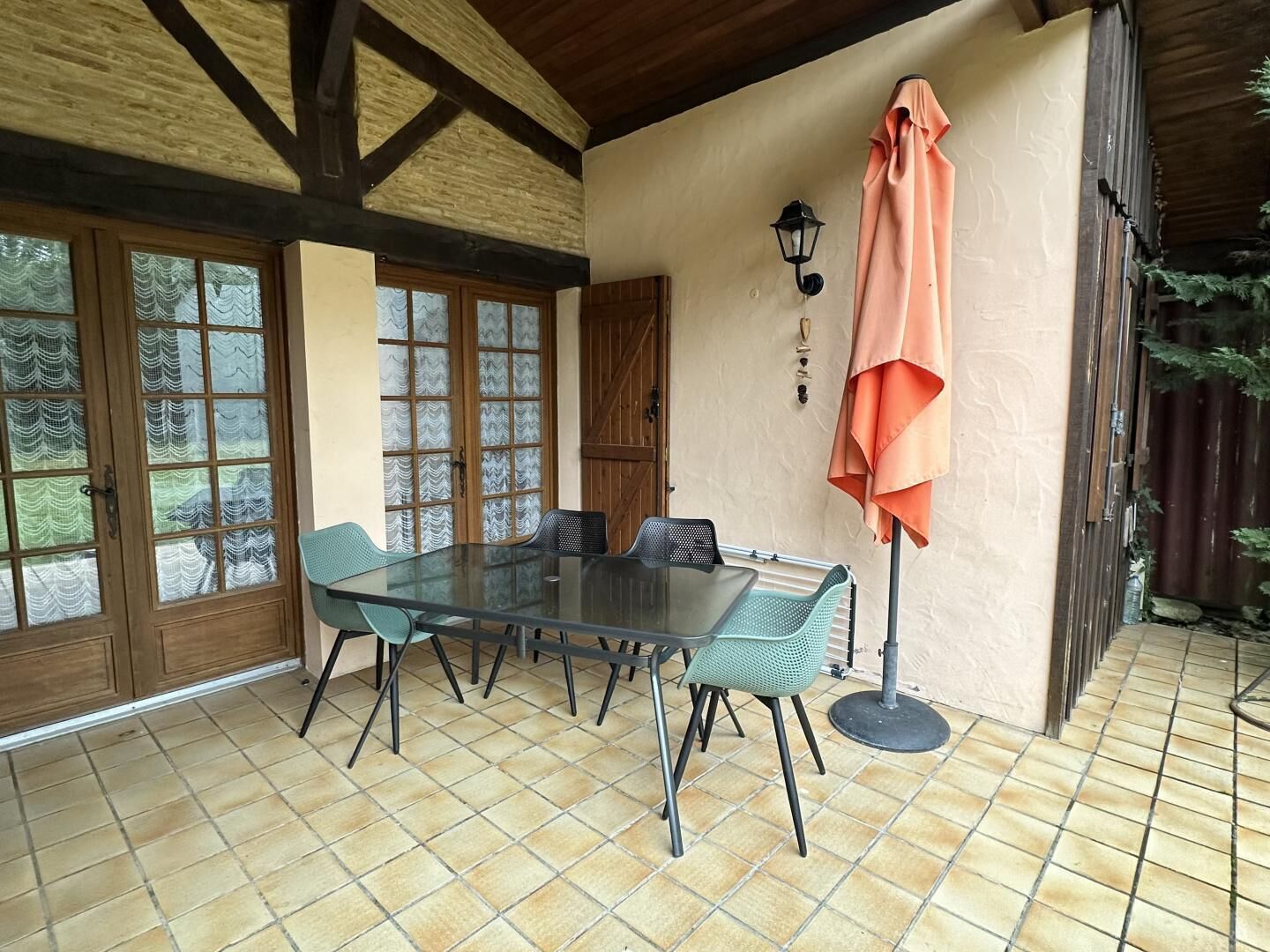 A covered patio with a glass table and chairs, wooden beams, a door with frosted glass panels, and a wall-mounted lamp.