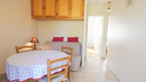 A compact room with a dining table, chairs, a bed with red pillows, wooden cabinets, and a lamp. - Chez Michel 1 - Angoume (ANGOUME)