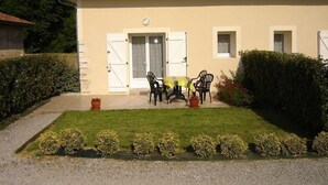 A house with a tiled roof, a front door, a small porch with a table and chairs, and a well-maintained lawn with hedges. - Chez Michel 1 - Angoume (ANGOUME)
