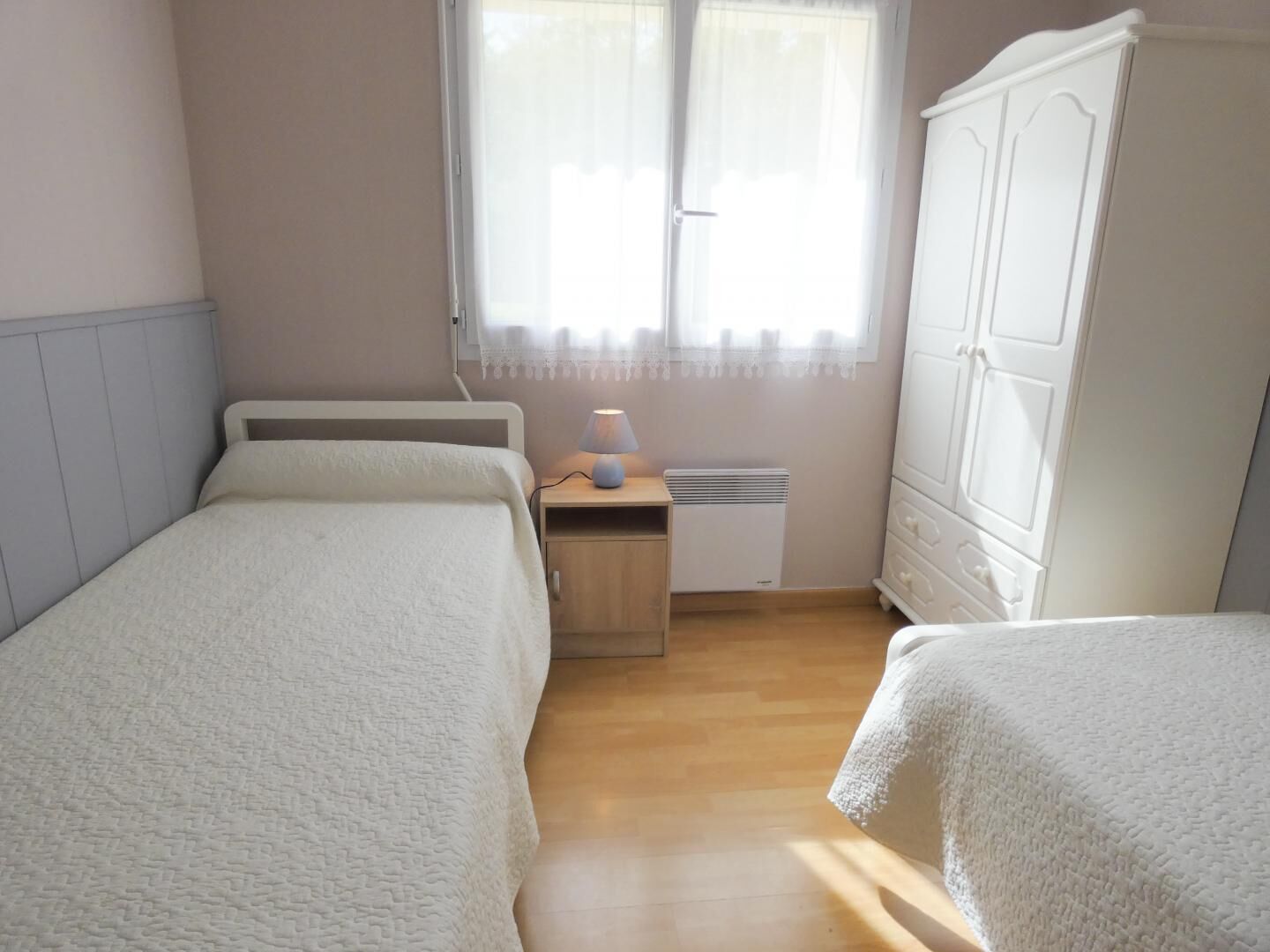 A bedroom with two beds, a nightstand with a lamp, a heater, and a wardrobe.