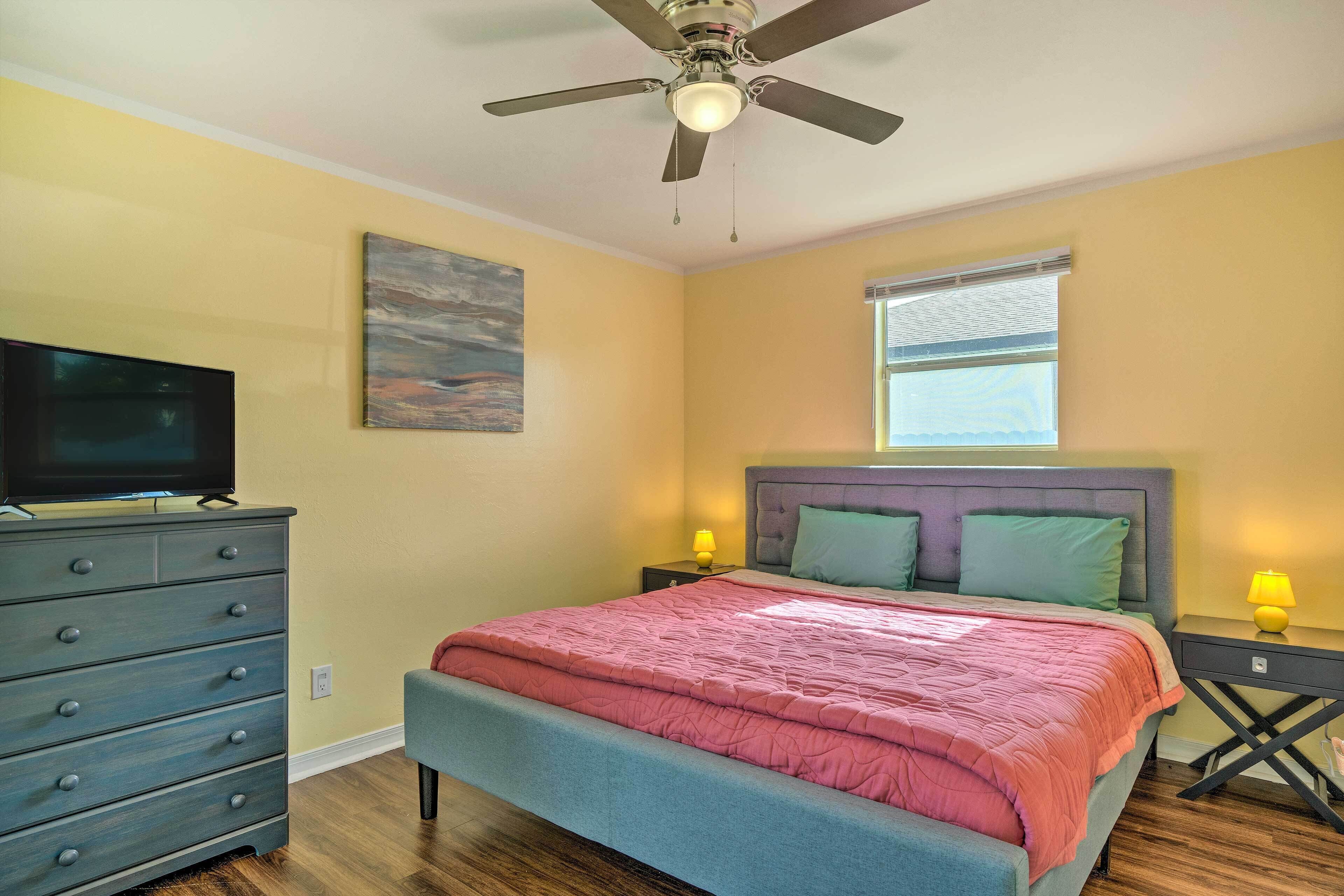 Une chambre à coucher avec un lit, une commode, un ventilateur de plafond, une fenêtre et un tableau accroché au mur.