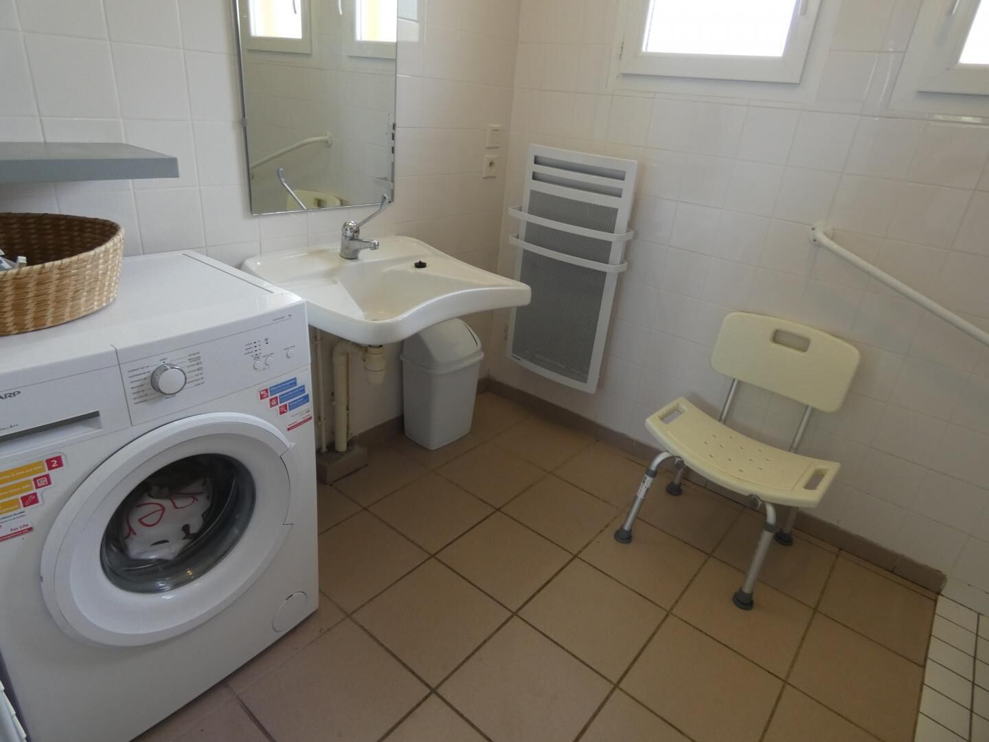 A compact bathroom with a washing machine, a sink, a small trash bin, a chair, and a window.
