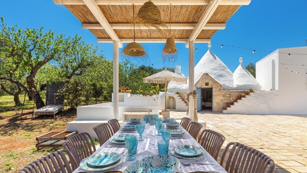 Ein Essbereich im Freien mit einem Tisch für sechs Personen, umgeben von Korbstühlen, unter einer Pergola mit hängenden Körben.
