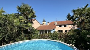 Une piscine entourée d’une végétation luxuriante et un bâtiment surmonté d’un toit recouvert de tuiles rouges.
