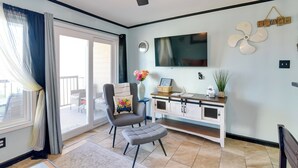 Una sala de estar con un sillón gris, un reposapiés a juego, un televisor montado en la pared, un ventilador y un ventanal corredizo que da a un espacio exterior.