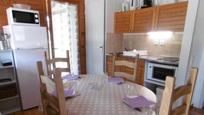 A compact kitchen with wooden cabinets, a microwave, a refrigerator, and a dining table set for two with chairs.