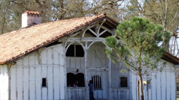 A rustic white building with a tiled roof, a porch with a railing, and a small tree in front.