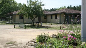 A rustic building with a tiled roof, surrounded by a garden with various flowers and a bench.